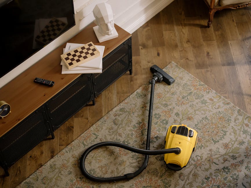 A yellow canister vacuum cleaner with a flexible hose and metal extension wand positioned on a patterned area rug in a living room, adjacent to a wooden media console with black metal legs. On top of 