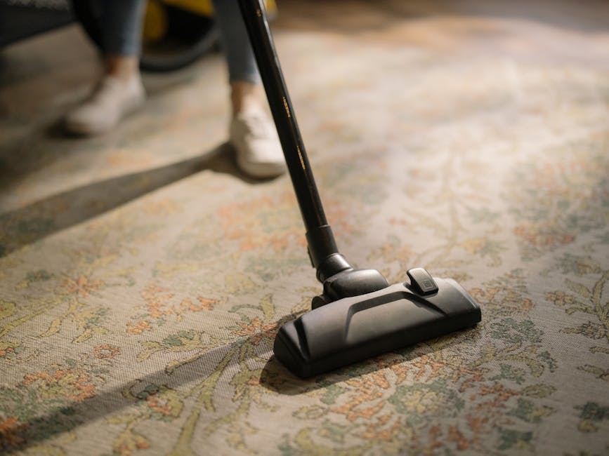 A yellow canister vacuum cleaner with a flexible hose and metal extension wand positioned on a patterned area rug in a living room, adjacent to a wooden media console with black metal legs. On top of 