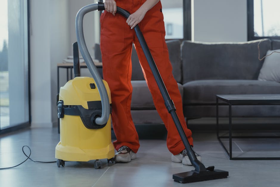 A close-up of a vacuum cleaner being used on a patterned area rug with floral motifs in a home setting. The vacuum head is black and rectangular, and the handle extends upward, held by a person wearin