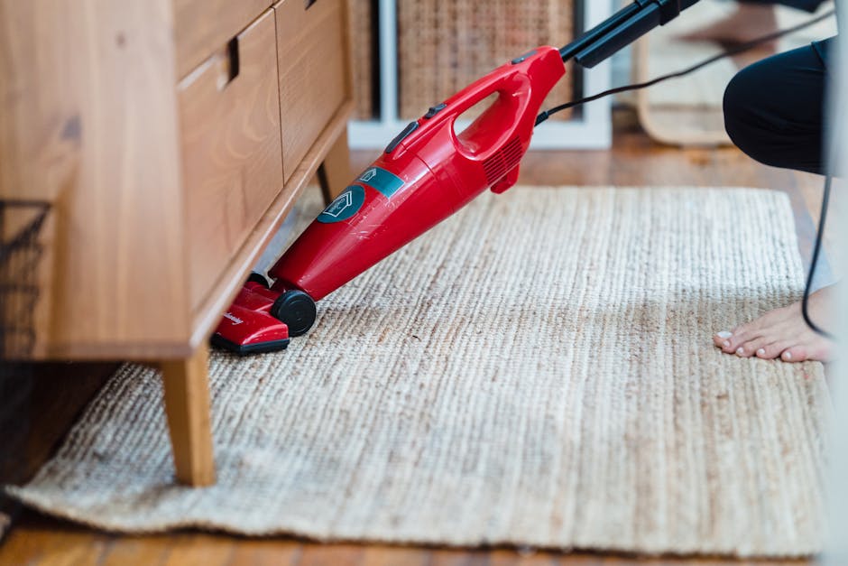 A person wearing beige shoes, blue jeans, and a beige jacket is using a vacuum cleaner to clean an ornate patterned area rug with floral motifs in shades of beige, green, and orange. The vacuum cleane