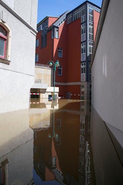 A flood-affected urban alleyway showing a large puddle of water reflecting the surrounding buildings, including a modern multi-story red-brick structure with glass windows and a white adjacent building. The alley is partially submerged, with the water reaching the base of a street lamp and covering the pavement. Bright sunlight illuminates the scene, emphasizing the water's surface and the contrast between the buildings' facades. This image highlights the impact of flooding on residential and commercial areas, illustrating the need for professional flood response and surface cleaning services offered by Carpet Cleaning Kingston for effective sanitisation and restoration.