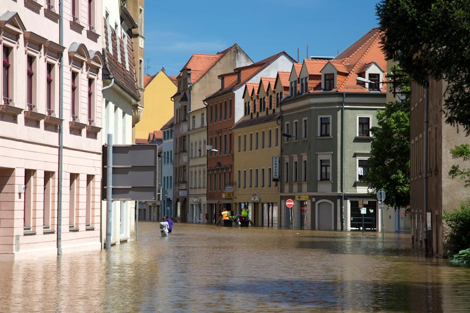 A flooded street in a residential area with colorful multi-story buildings on either side, featuring varied architectural styles and red-tiled roofs. The water covers the entire road surface, partially submerging the lower floors of the buildings and a parked car. Some pedestrians are seen wading through the floodwaters, while others are standing on the pavement. The bright blue sky and sunlight highlight the wet surfaces, and a large tree with dense foliage is visible on the right side. This image depicts the aftermath of flooding in Kingston, emphasizing the urgent need for emergency water removal and surface cleaning by professionals such as Carpet Cleaning Kingston.