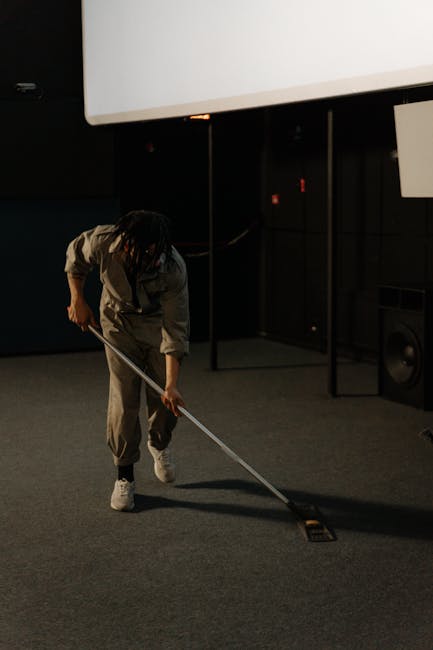 A woman with light-colored hair, dressed in a casual top and overalls, is performing surface cleaning using a vacuum cleaner with a hose attachment on a tiled floor. The room features large glass slid