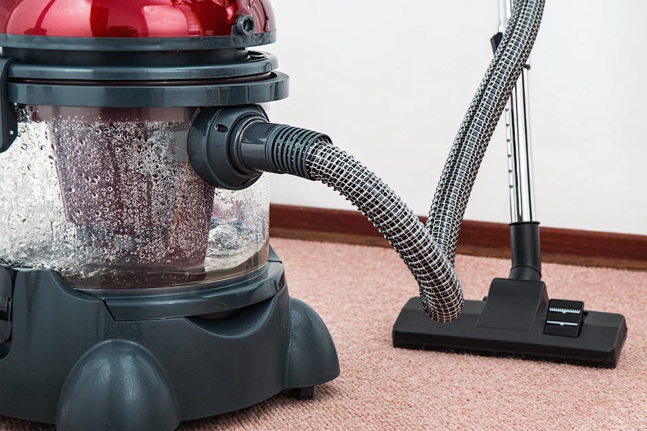 A professional cleaner dressed in full white protective suit, including gloves, mask, and shoe covers, is performing deep carpet cleaning in a bright, modern living room. The cleaner is using a handheld vacuum or extraction machine on a plush, beige carpet, which appears to be wet and soiled before treatment. The room features white walls, a large window allowing natural light, and potted plants near the window for decor. There is a small wooden side table and a shelving unit with decorative items and storage boxes. The scene exemplifies comprehensive domestic cleaning, focusing on surface sanitation and renewal of the carpet surface, consistent with professional cleaning standards offered by Carpet Cleaning Kingston as seen on the webpage for same-day cleaning in Surbiton High Street residents KT6.