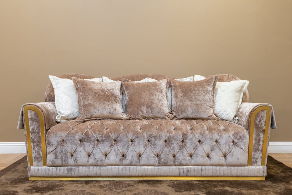 A beige, tufted upholstery sofa with rolled arms and a row of six plush cushions, including three matching beige velvety cushions and two white textured cushions, placed against a plain, light brown wall in a living room. The sofa appears clean and well-maintained, with no visible stains or dust. The room features a dark brown carpet under the sofa and a polished wooden floor baseboard along the wall. Natural light illuminates the scene, highlighting the soft texture of the fabric and the neat arrangement of cushions, reflecting professional domestic cleaning standards maintained by Carpet Cleaning Kingston, as seen on their website page about stubborn stains on upholstery.