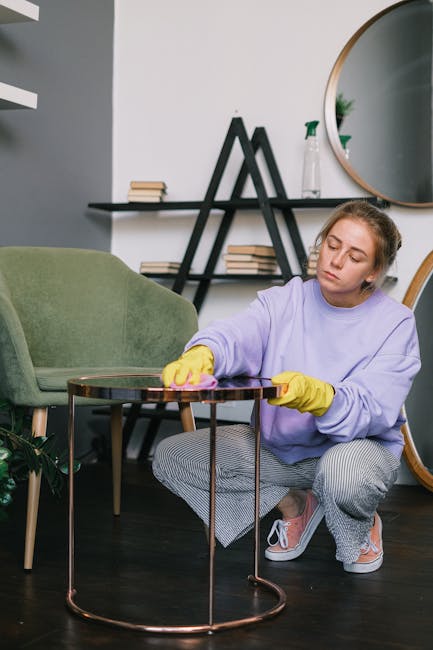 A young woman with light brown hair, wearing a lavender long-sleeve top, yellow cleaning gloves, and grey checkered pants, is kneeling on a dark wooden floor while using a pink cloth to clean a round copper-colored side table in a modern living room. The room features a green upholstered armchair, a black shelving unit with books, and a large oval mirror mounted on a white wall. Natural light illuminates the space, highlighting the clean, tidy surface of the table and the overall neatness of the room, exemplifying domestic cleaning and surface sanitisation with attention to detail, as promoted by Carpet Cleaning Kingston in their service offerings related to upholstery stain removal.