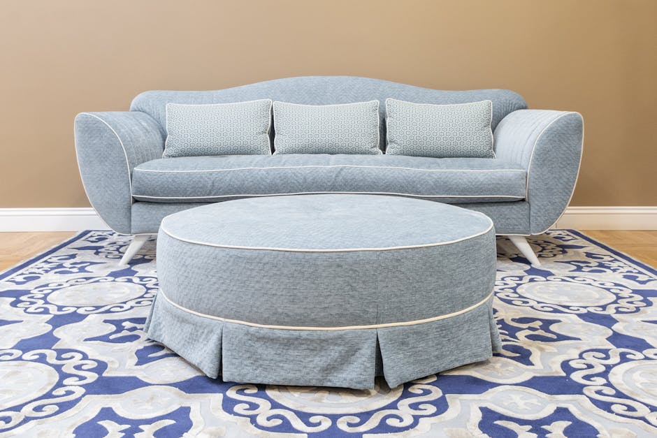 A beige, tufted upholstery sofa with rolled arms and a row of six plush cushions, including three matching beige velvety cushions and two white textured cushions, placed against a plain, light brown wall in a living room. The sofa appears clean and well-maintained, with no visible stains or dust. The room features a dark brown carpet under the sofa and a polished wooden floor baseboard along the wall. Natural light illuminates the scene, highlighting the soft texture of the fabric and the neat arrangement of cushions, reflecting professional domestic cleaning standards maintained by Carpet Cleaning Kingston, as seen on their website page about stubborn stains on upholstery.